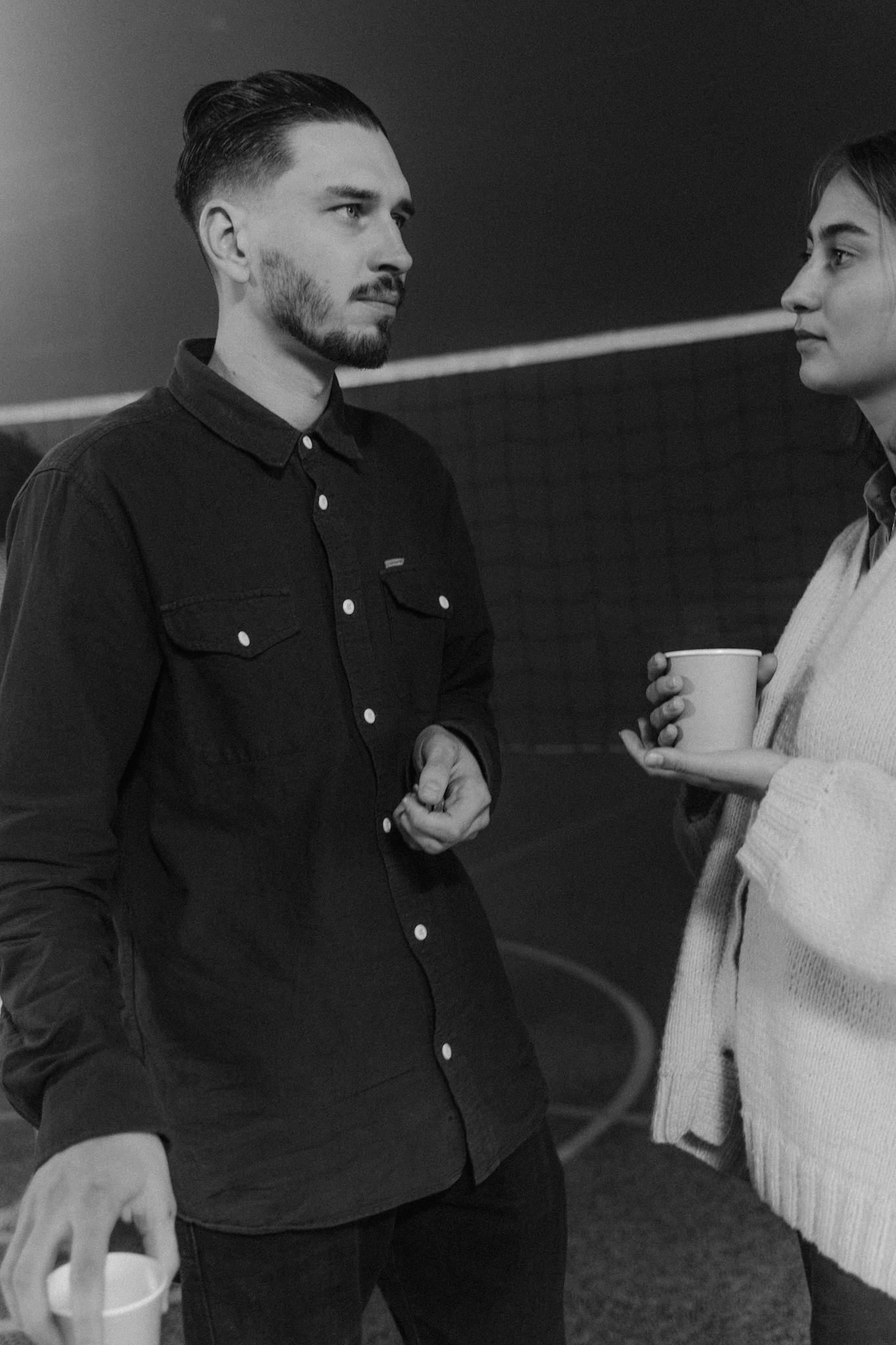 Two adults talking in a moody, dimly-lit indoor space, holding cups.